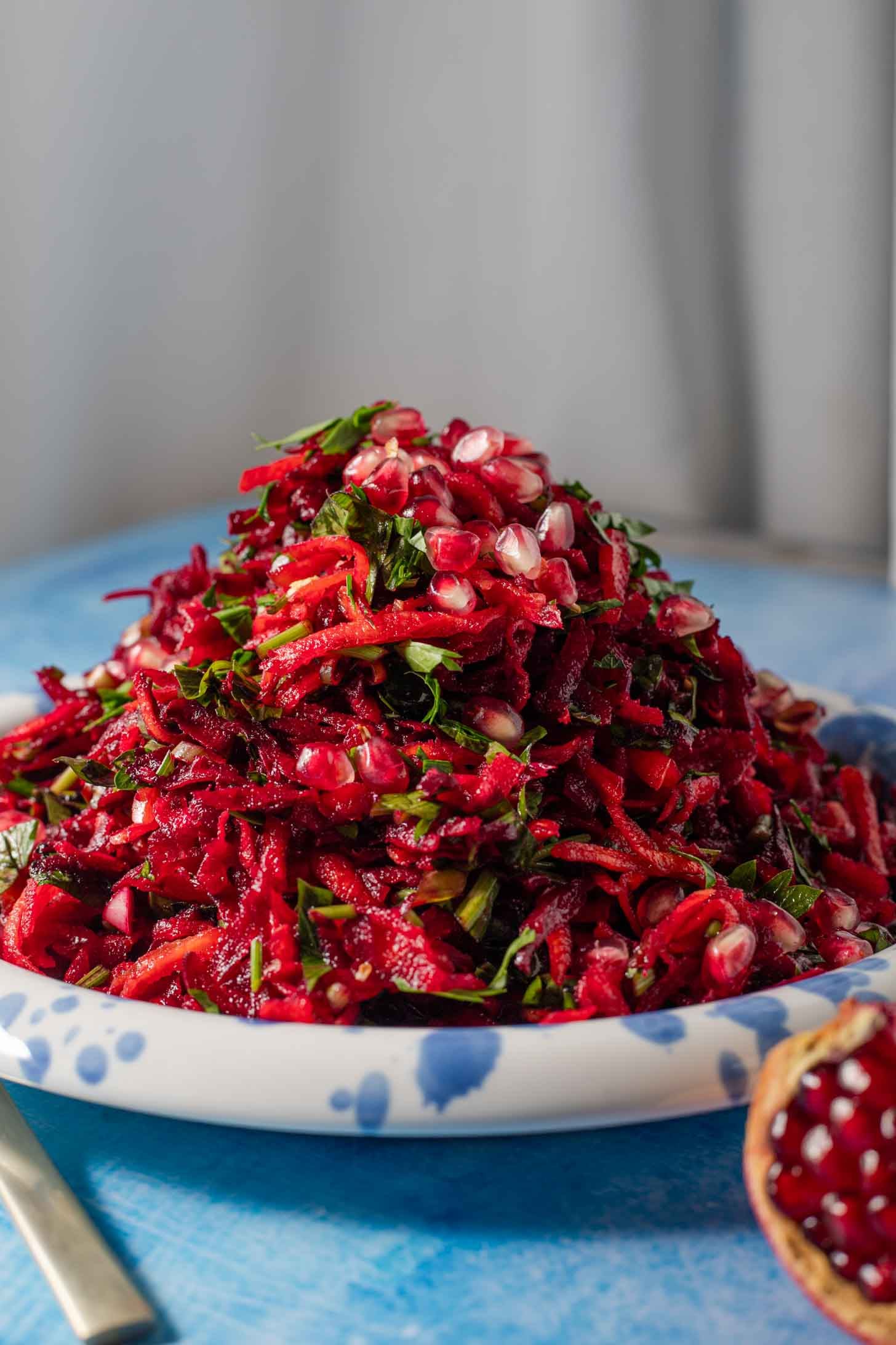 A plate piled with shredded beet salad, garnished with fresh herbs and pomegranate seeds, sits on a blue surface. A pomegranate half is visible in the foreground.