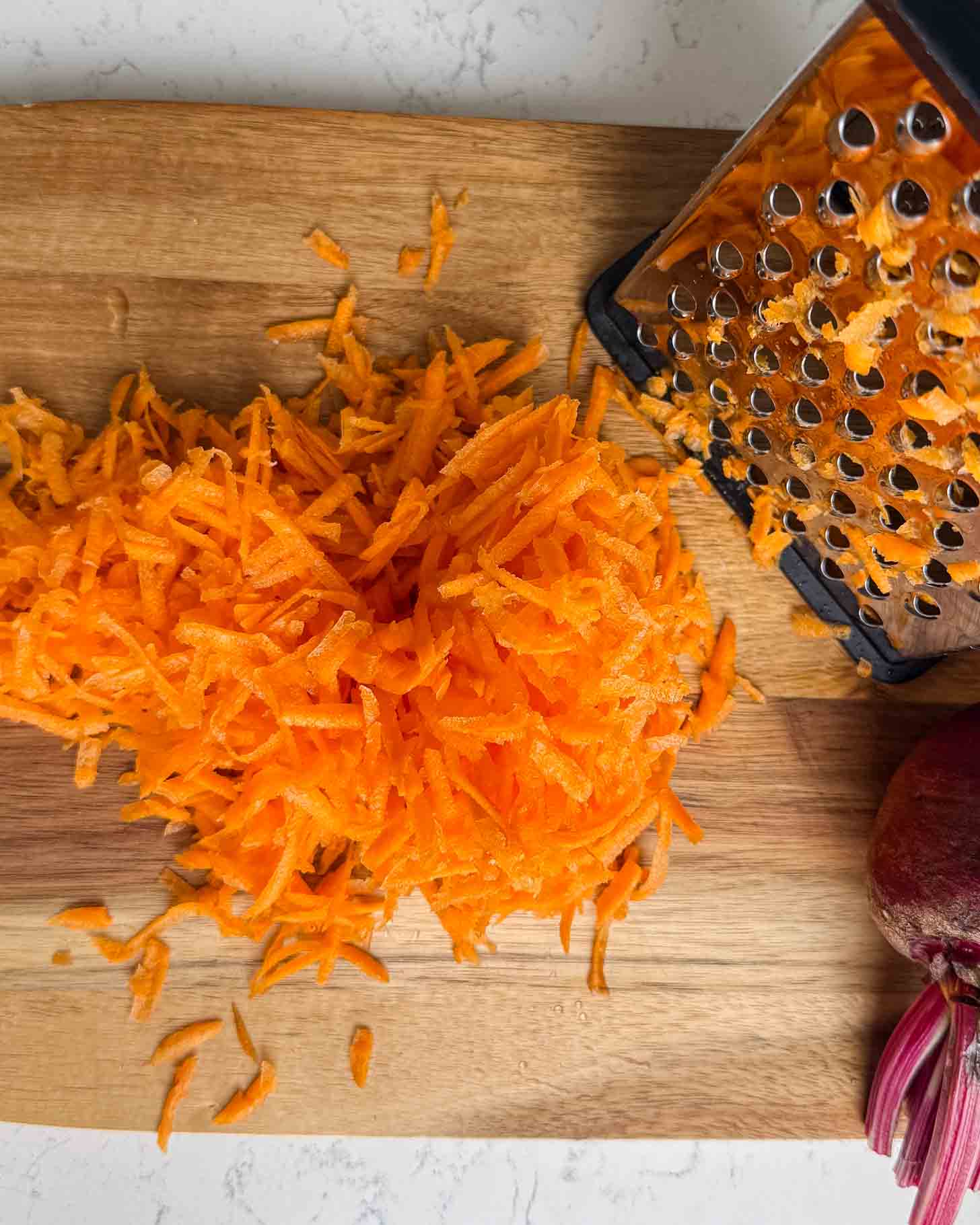 A pile of freshly grated carrots on a wooden cutting board, next to a metal box grater and part of a red onion in the corner.
