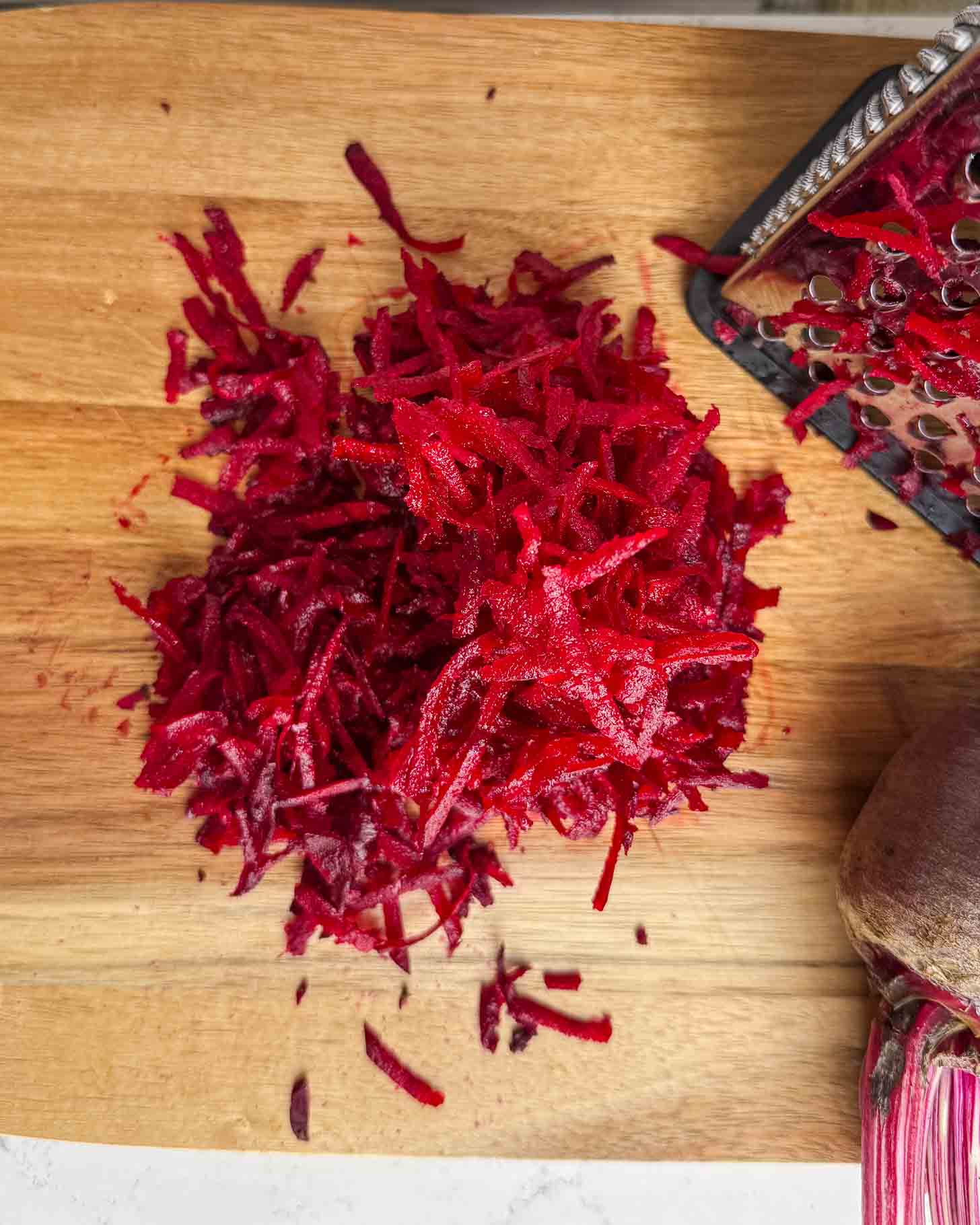 A pile of freshly grated beetroot sits on a wooden cutting board next to a metal grater and part of a whole beetroot.