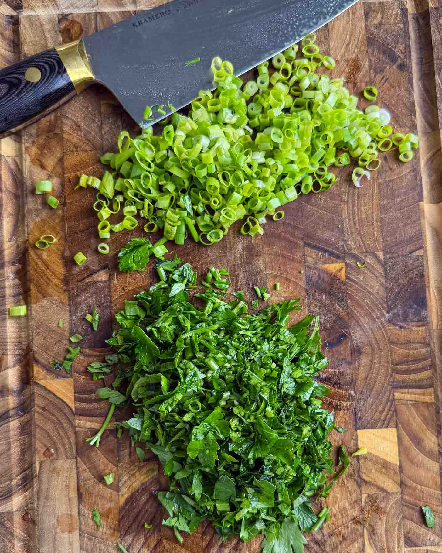A knife rests on a wooden cutting board with chopped green onions and chopped parsley arranged in separate piles.