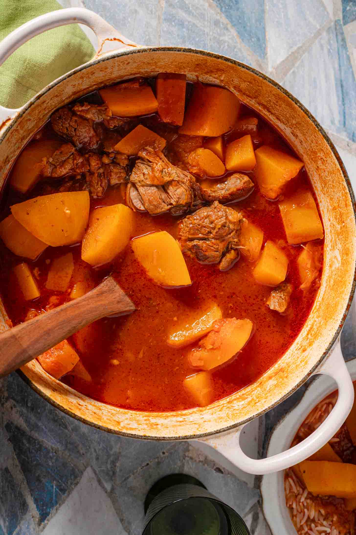 A white enameled Dutch oven filled with beef and potato stew in a reddish broth, with a wooden spoon resting inside.