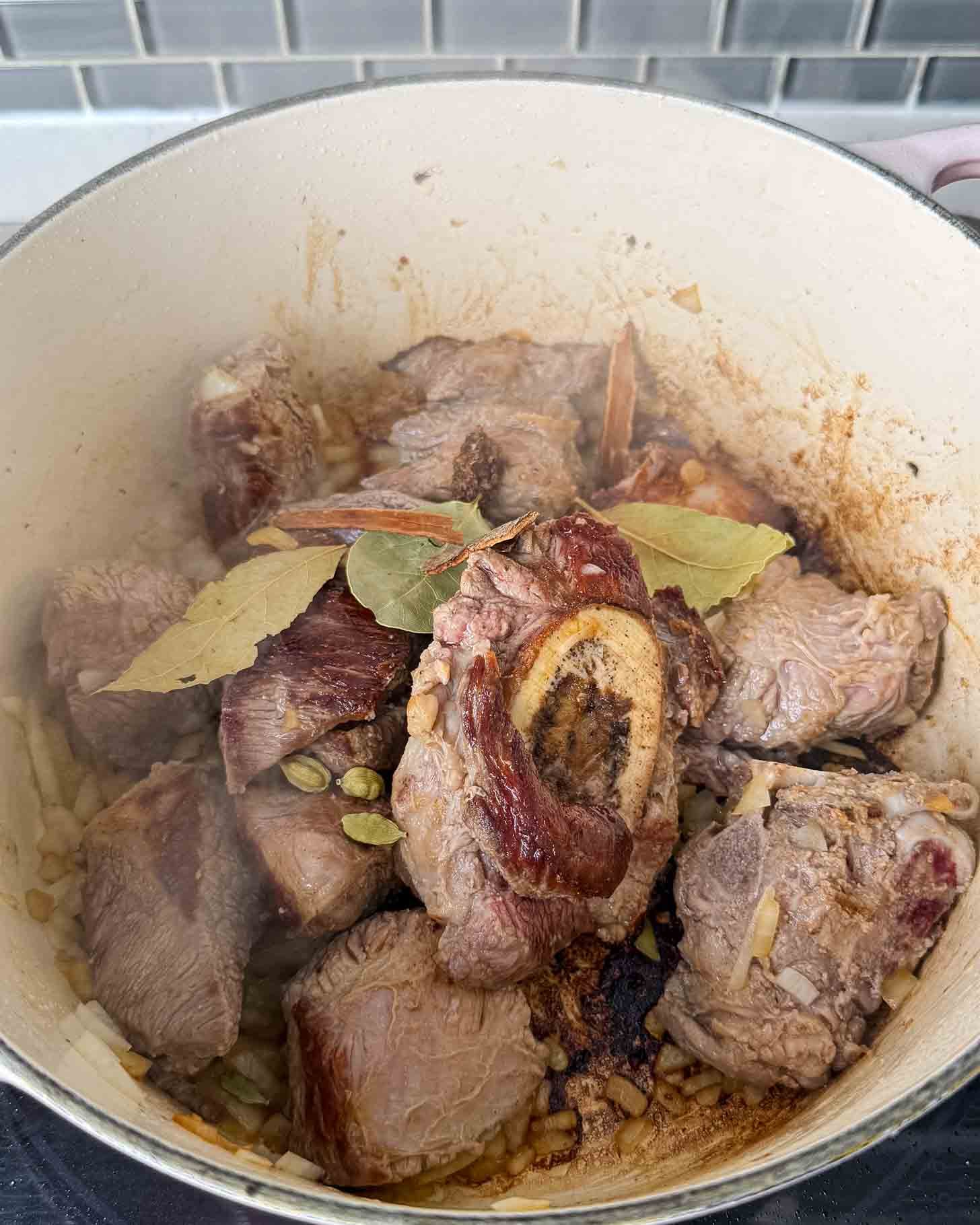 Chunks of beef, including a bone-in piece, searing in a pot with bay leaves, cardamom pods, and cinnamon sticks.