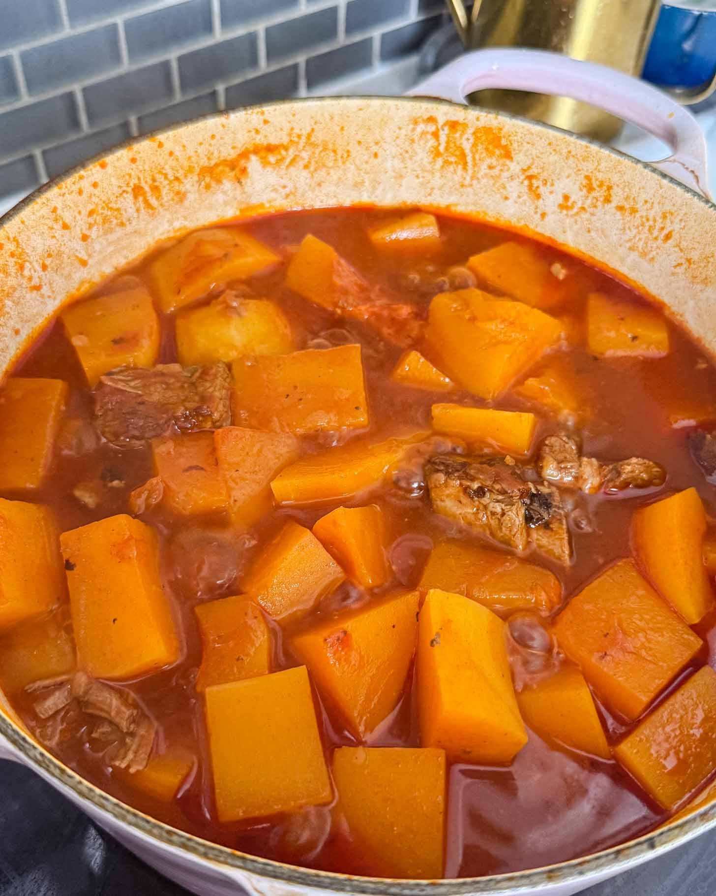 A stew of cubed orange squash and tender meat simmering in a red tomato-based broth inside a dutch oven.
