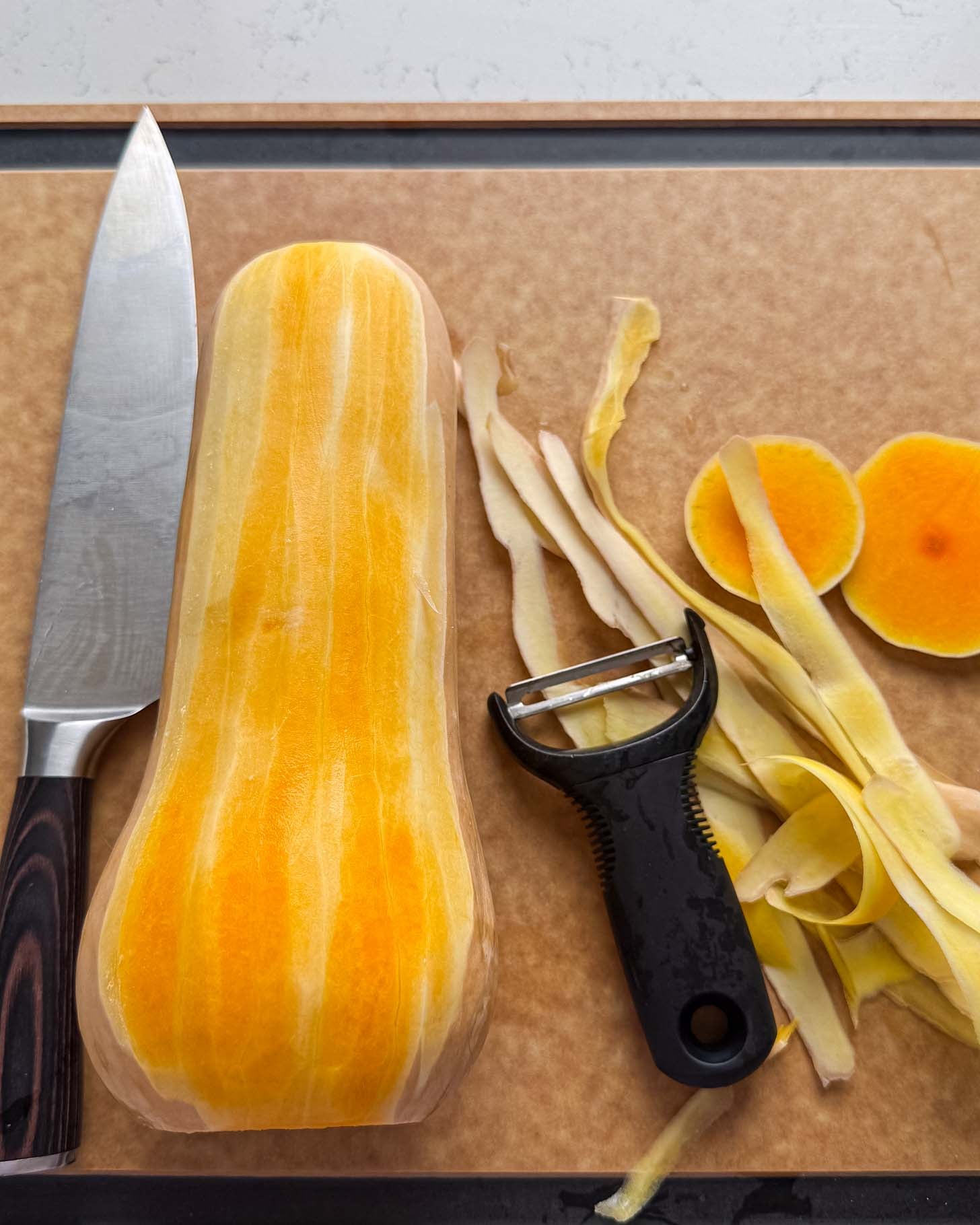 A peeled butternut squash on a cutting board with a chefs knife, a vegetable peeler, and squash peelings.