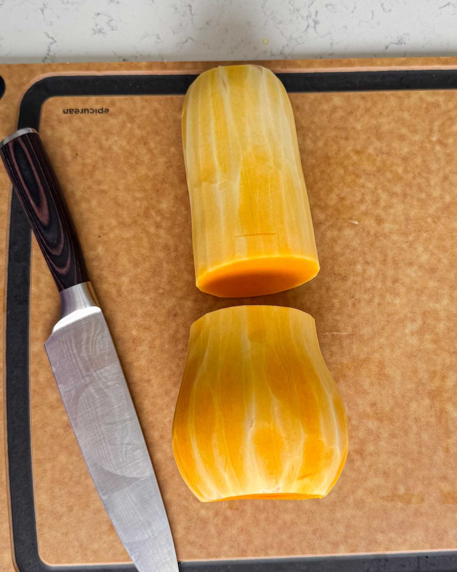 A peeled butternut squash cut into two pieces on a cutting board next to a chefs knife.