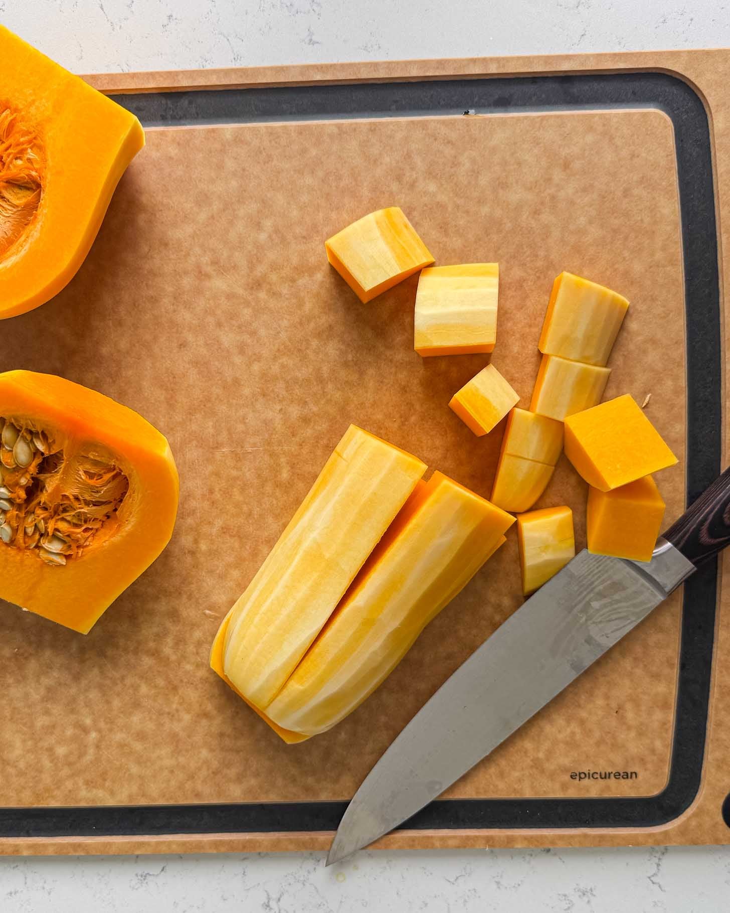 A butternut squash cut into halves and cubes on a cutting board next to a chefs knife.