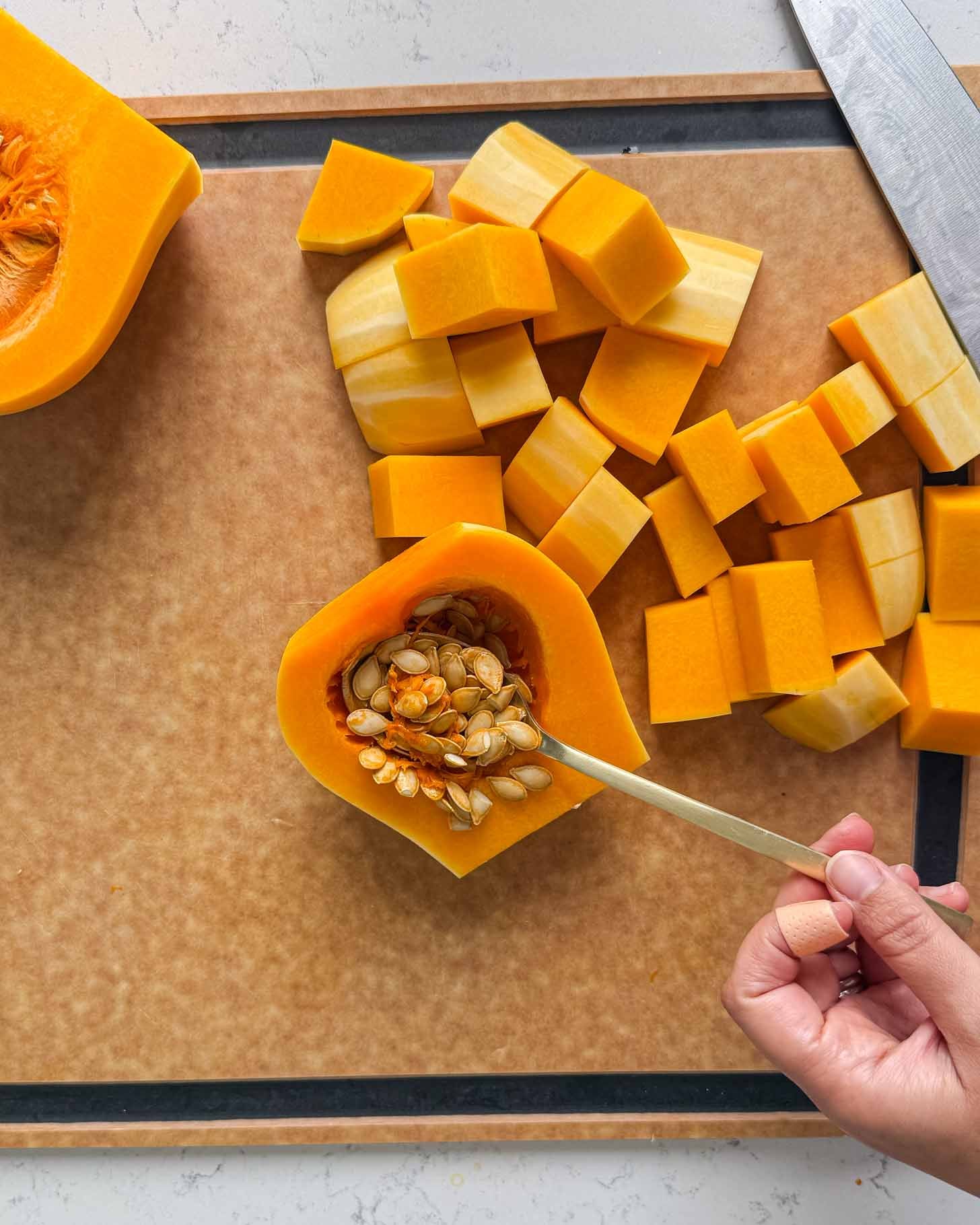 A hand uses a spoon to scoop seeds out of a halved butternut squash on a cutting board, surrounded by cubes of peeled squash and a kitchen knife.