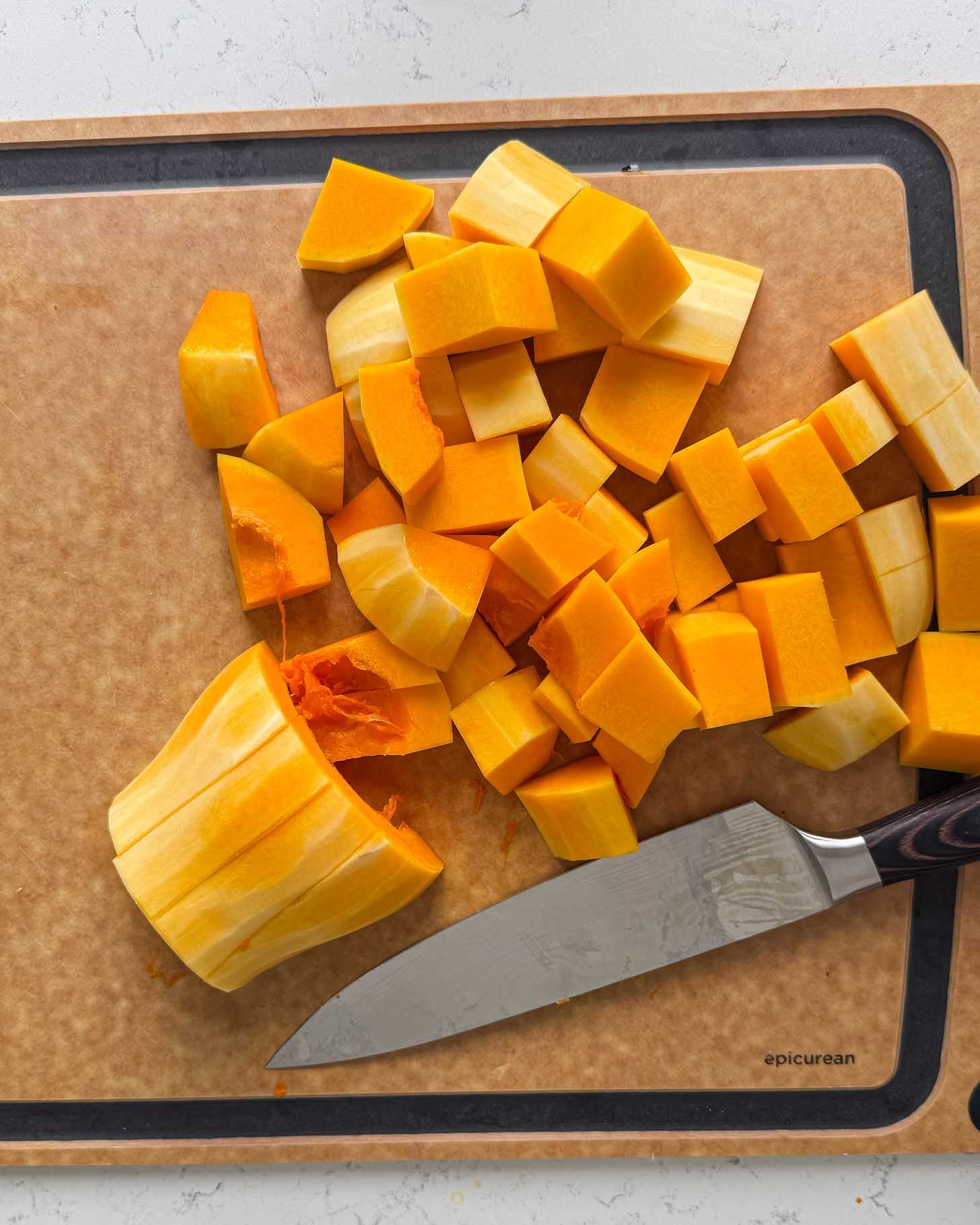 A large piece and many smaller cubes of peeled butternut squash on a cutting board next to a chefs knife.
