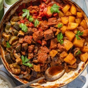 A large skillet containing a stew of cubed meat, potatoes, and vegetables, garnished with fresh parsley and served with a side of white rice.
