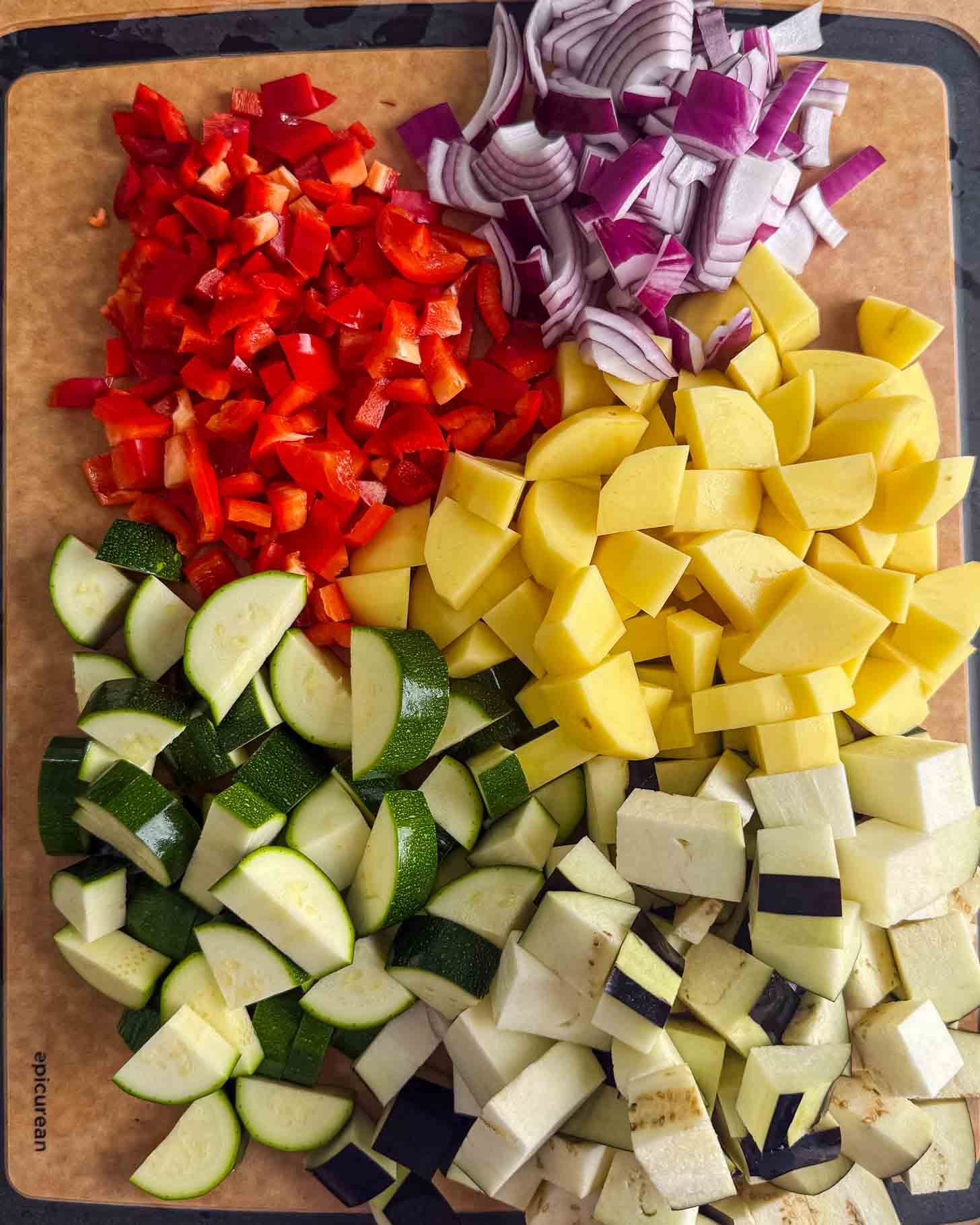 A cutting board topped with piles of chopped red bell peppers, red onion, yellow potatoes, zucchini, and eggplant.