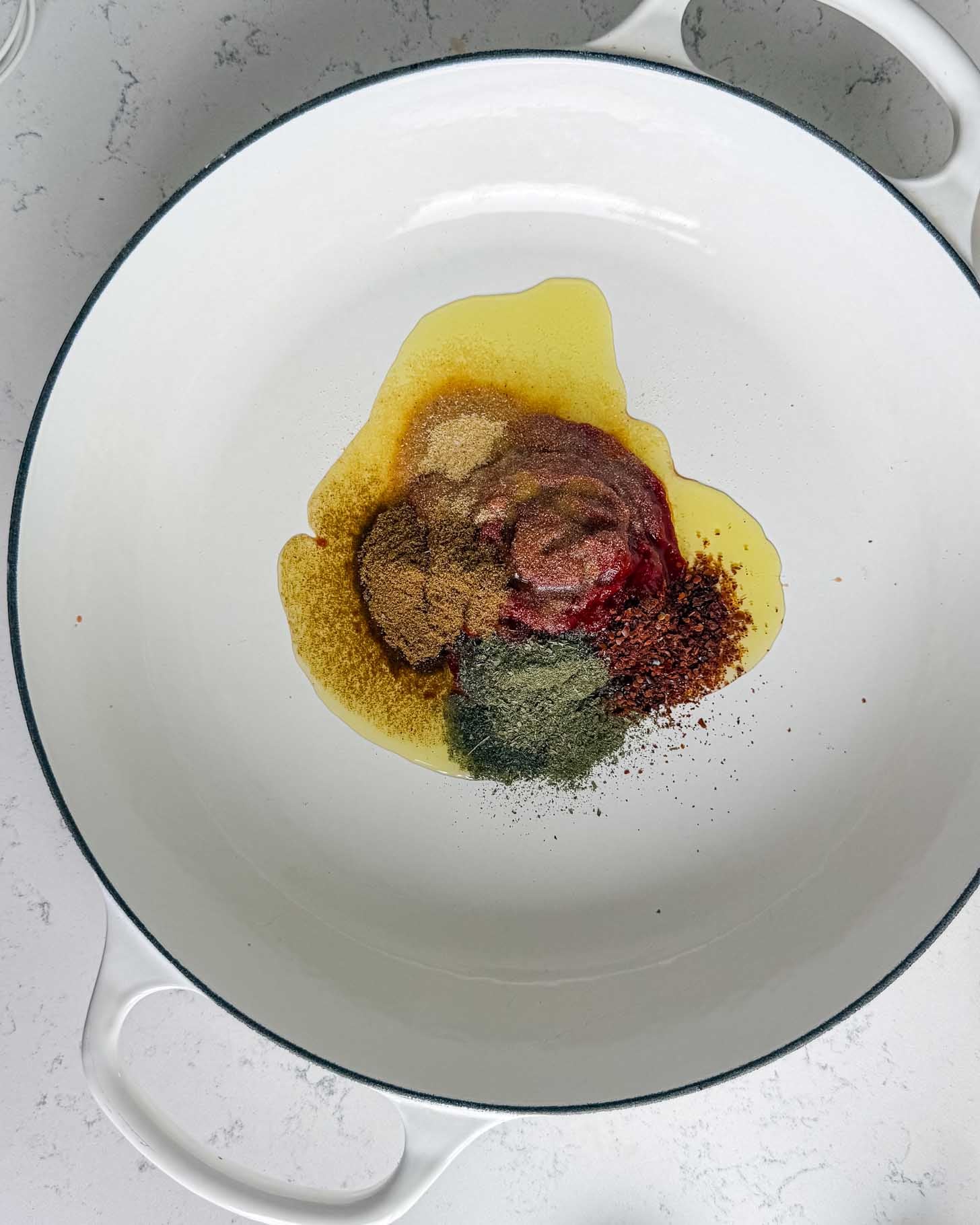 A pool of olive oil in a white enameled pan topped with piles of tomato paste and various ground spices.