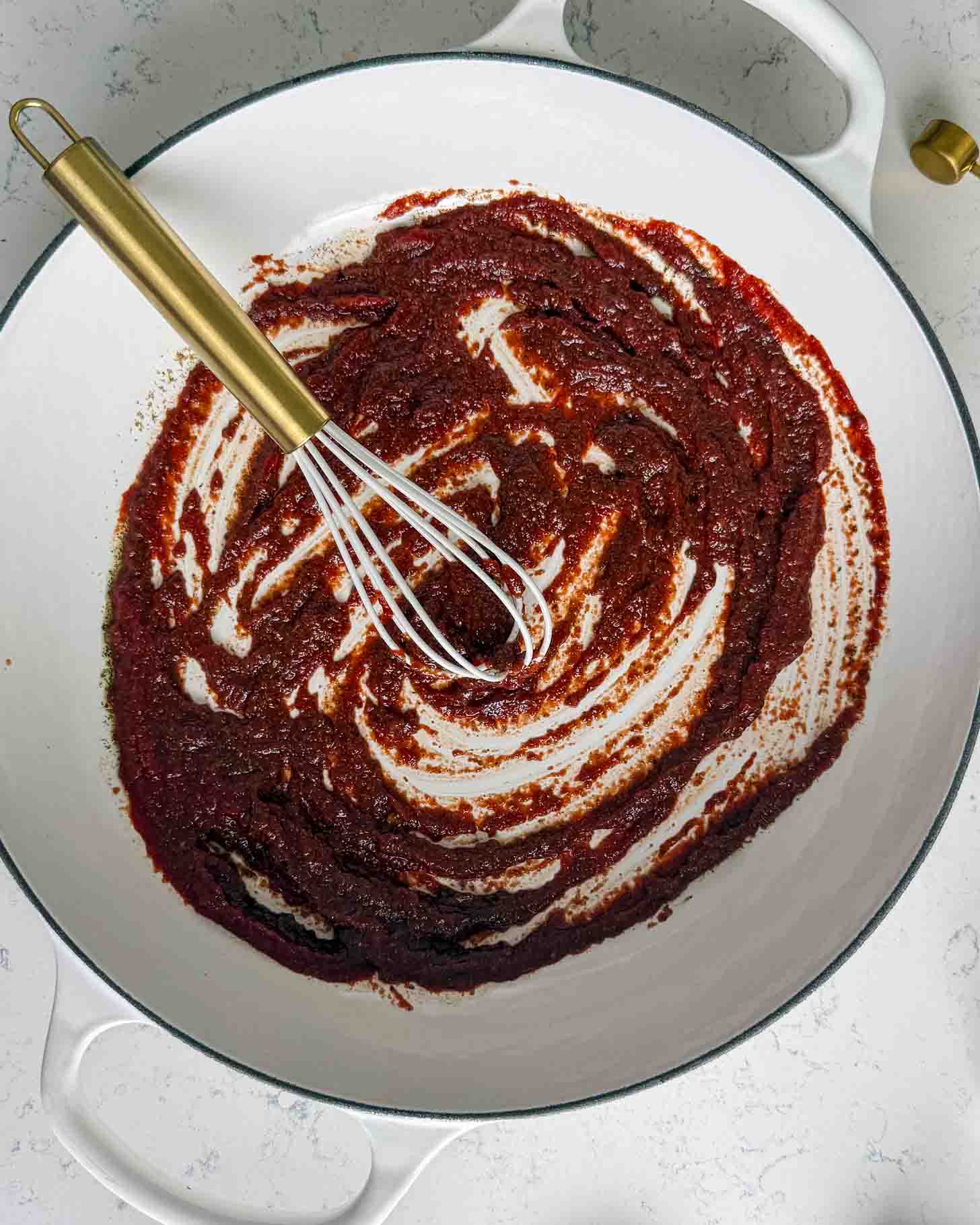A whisk mixing a thick, dark red sauce in a white enameled skillet.