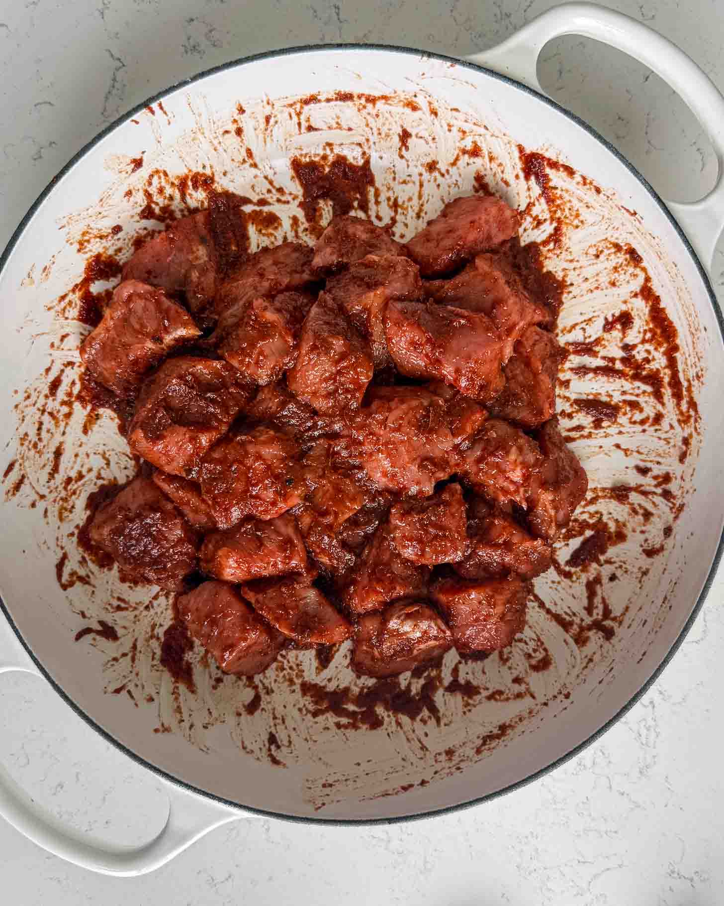 Cubes of raw meat coated in a thick red sauce in a white enameled cast iron pot.