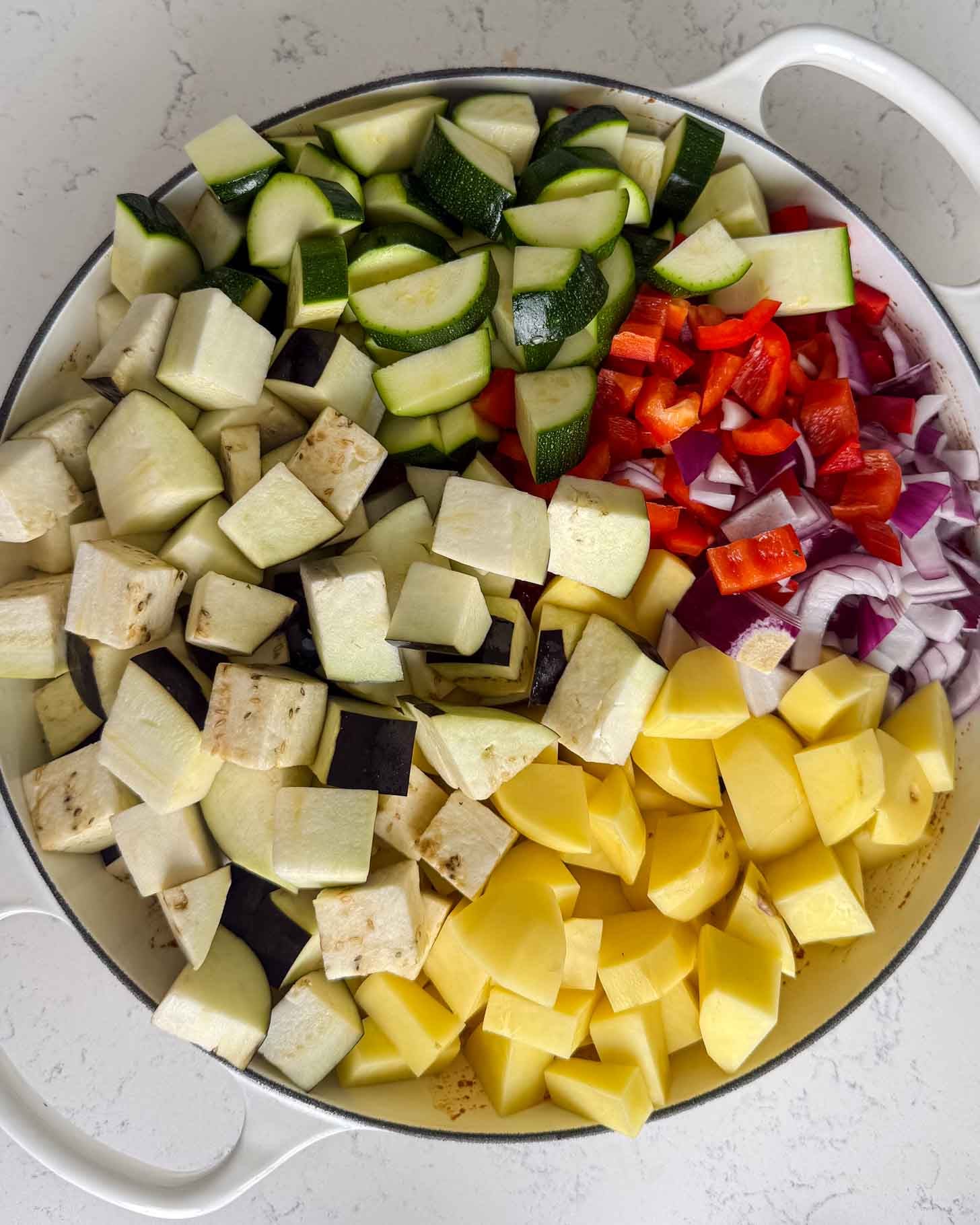 A large pan filled with chopped zucchini, eggplant, potatoes, red bell peppers, and red onions.