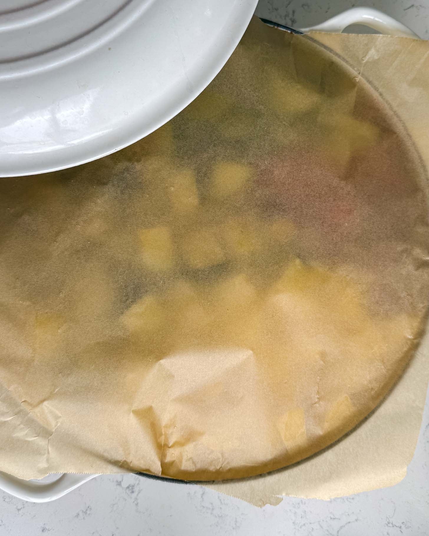 A round baking dish filled with chopped produce, covered with a sheet of parchment paper and a partially lifted white lid.